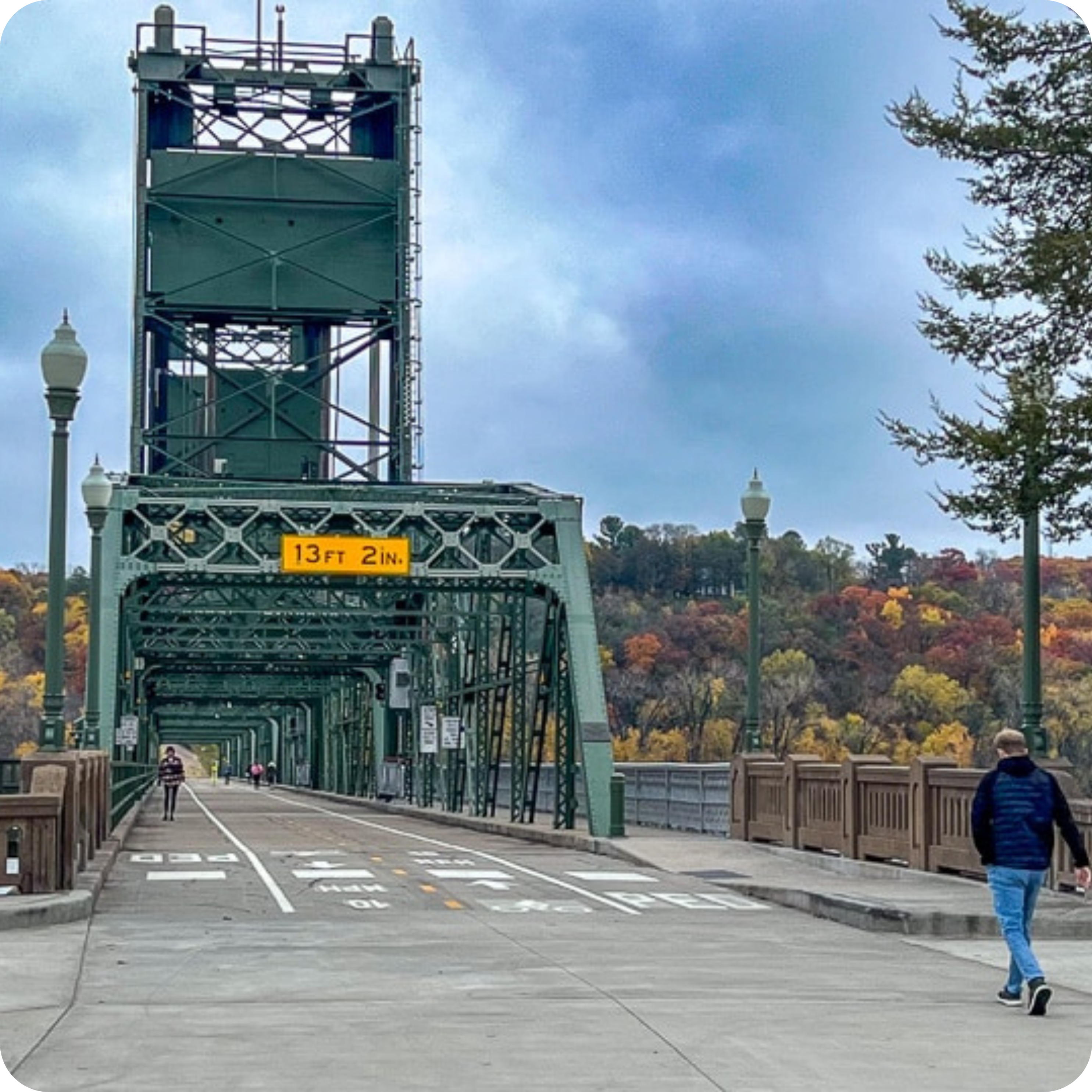 A green lift bridge in Stillwater, MN, featuring two individuals—one walking and one cycling—under an overcast sky, with an autumn landscape in the background. A height restriction sign displays 